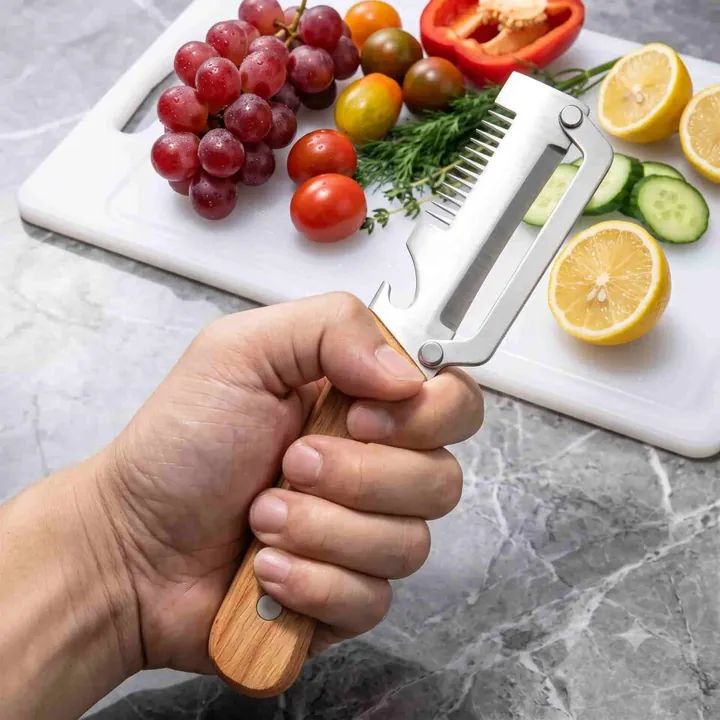 A hand holds the PeelPro peeler over a cutting board with tomatoes, grapes and a lemon